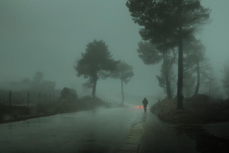 A lone person walking on the side of the road in the rain. The taillights of a car is visible in the background.